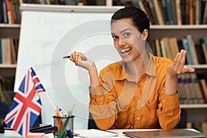 Online foreign languages tutoring. Friendly female teacher giving english class, sitting near blackboard in library