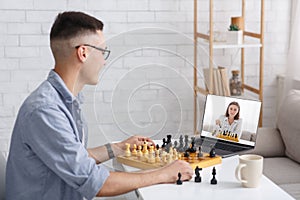 Online game with rival and modern gadgets. Smiling guy plays board game and looks at laptop at lady opponent with chess