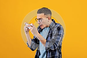 Online gaming. Addicted young guy playing video games on cellphone, overusing his gadget on orange studio background