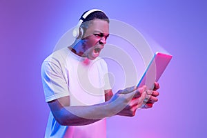 Online Gaming. Angry African Man In Wireless Headphones Using Digital Tablet