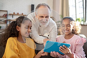 Grey-haired man in a white shirt lloking at the tablet together with two girls photo