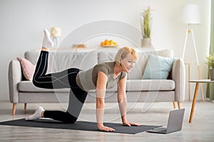 Online home fitness concept. Beautiful mature woman exercising to sports video on laptop in living room