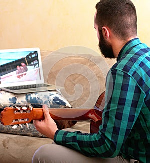 Online learning concept. Young man concentrating as he learns to play the guitar thanks to a tutorial on his laptop at home