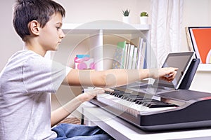 Boy learning music online. Child watching video tutorial at tablet computer and playing digital piano at home