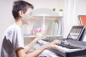 Boy watching video lesson at tablet computer and having fun playing piano at home. Online learning remote education