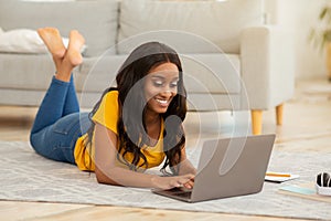 Online life concept. Gorgeous black woman lying on floor with laptop computer, working or studying from home