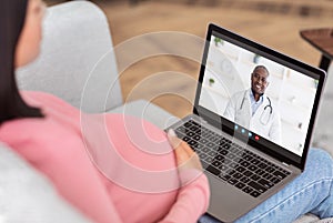Online medical consultation. Expectant woman having video call with friendly black doctor on laptop from home