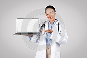 Online medical health support. Cheerful latin female doctor with stethoscope showing blank screen on laptop, mockup