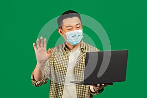 Online meeting concept. Asian mature man in surgical face mask making video call on laptop, waving at computer webcamera