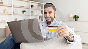 Online payment concept. Happy guy using laptop and credit card, sitting on comfy sofa, panorama, copy space