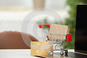 Online payment concept. Small shopping cart with bank card, box and laptop on white table