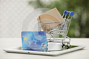 Online payment concept. Small shopping cart with bank card, box and tablet on white table, closeup