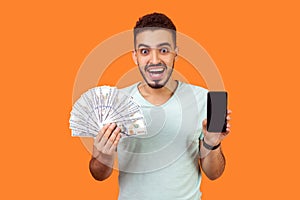 Online payment. Portrait of extremely happy man holding money and cellphone. indoor studio shot isolated on orange background