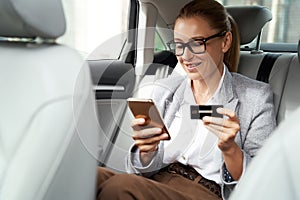 Online payment. Smiling business woman wearing eyeglasses using her smartphone and credit card to buy something while