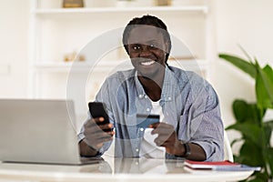 Online Payments. Smiling Black Male Freelancer Using Smartphone And Credit Card