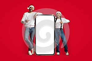 Online Sale. Joyful Couple Wearing Santa Hats Pointing At Big Blank Smartphone