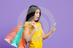 Online Sales. Shocked Asian Woman Looking At Smartphone And Holding Shopping Bags