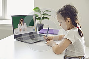 Online school education. A little girl a schoolgirl writes in a notebook has a video lesson with a female laptop teacher