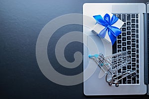 Online shopping background. Laptop computer, shopping trolley and white gift with blue ribbon on dark. Internet purchase