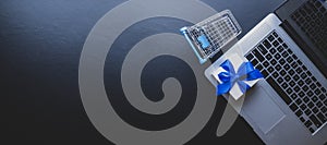 Online shopping background. Laptop computer, shopping trolley and white gift with blue ribbon on dark. Internet purchase