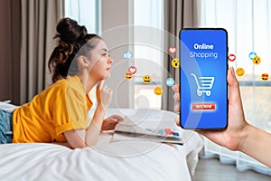 Online shopping. Blurred young woman is lying on the bed with magazine. Hand with smartphone with grocery cart and flying emoji.