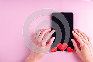 Online shopping concept. Female hands on a black blank tablet screen with red heart shape on a pink background