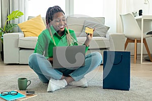 Online shopping concept. Happy black woman showing credit card and using laptop, ordering clothes via internet