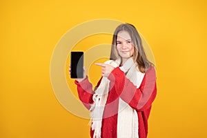 Online shopping concept. Smilling teen girl showing empty smartphone screen and pointing on it, yellow background