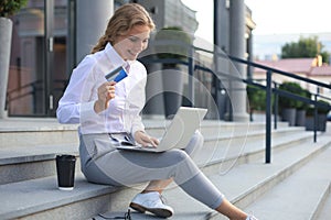 Online shopping concept. Young blonde woman holding a credit card and doing online payment with laptop outdoors