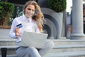 Online shopping concept. Young blonde woman holding a credit card and doing online payment with laptop outdoors