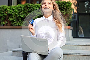 Online shopping concept. Young blonde woman holding a credit card and doing online payment with laptop outdoors