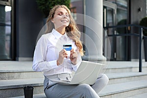 Online shopping concept. Young blonde woman holding a credit card and doing online payment with laptop outdoors