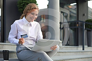 Online shopping concept. Young blonde woman holding a credit card and doing online payment with laptop outdoors