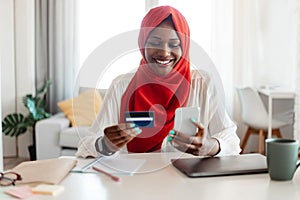 Online shopping and e-payment. Happy muslim woman in hijab using cellphone and credit card, sitting at workplace at home