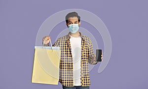 Online shopping during epidemic. Young Indian guy holding bag and smartphone on lilac background, mockup for design