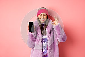 Online shopping and fashion concept. Fashionable senior asian woman showing blank black smartphone screen and OK sign