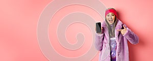 Online shopping and fashion concept. Stylish asian senior woman in faux fur coat pointing finger at blank smartphone