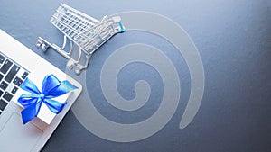 Online shopping flat. Laptop computer, shopping trolley and white gift with blue ribbon on dark background. Internet