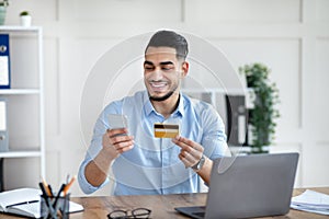 Online shopping. Happy Arab businessman holding credit card and smartphone, making e-payment, ordering goods on web