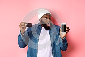 Online shopping. Impressed and surprised Black man showing credit card, staring at smartphone screen, demonstrate app