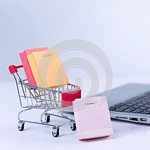 Online shopping. Mini shop cart trolley with colorful paper bags over a laptop computer on white table background, buying at home