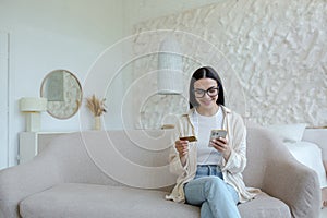 Online shopping. Satisfied young beautiful woman in glasses sitting on sofa at home. Holds a credit card and phone, pays