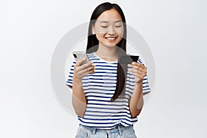 Online shopping. Smiling asian girl using mobile phone and credit card, paying in smartphone, standing over white