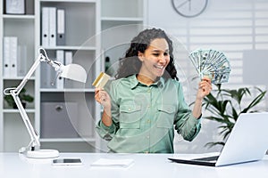 Online shopping. A young Arab woman is sitting in the office at a table with a laptop, holding a credit card and cash
