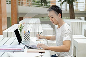 Online studying concept a male student in white t-shirt studying online by using his new white laptop and the calculator in the