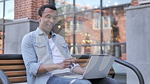 Online video chat on laptop by african man sitting on bench