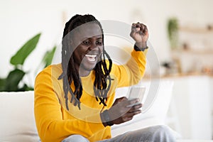 Online Win. Overjoyed Black Man Celebrating Success With Smartphone At Home