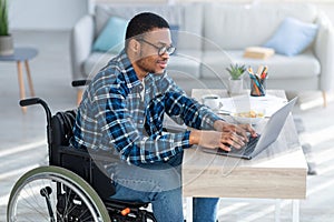 Online work for disabled people. Handicapped black guy in wheelchair using laptop for remote job at home