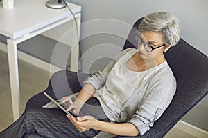 Online work education communication. Senior woman with glasses uses tablet at home.