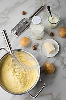 Ooking pot with mashed potatoes and ingredients as potato, milk, salt, butter, nutmeg with whisker and grinder on light marble